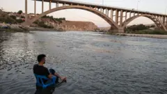 Homem sentado com cadeira de plástico na margem do rio Tigre, sobre o qual passa também uma grande ponte que pode ser vista