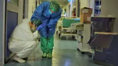 Em corredor de hospital, um membro da equipe consola outro, ajoelhado com as mãos na cabeça