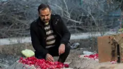Homem se agacha em frente a túmulo com flores