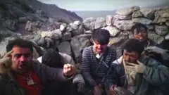 Grupo de hombres con cadenas al cuello sentados junto a un muro en la cima de una montaña