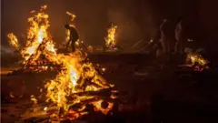 Workers and relatives stand around burning funeral pyres at night