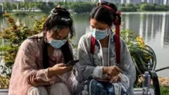 Two Chinese girls dressed in traditional ceremonial clothes look at their phones in the city of Wuhan