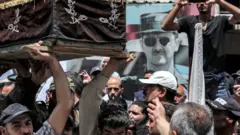 صورة للرئيس السوري بشار الأسد تلوح في الأفق على الحائط بينما يحمل المشيعون نعوش القتلى من أعضاء الجبهة الشعبية لتحرير فلسطين - القيادة العامة خلال جنازتهم بعد عودتهم إلى وطنهم في مخيم اليرموك للاجئين الفلسطينيين جنوب دمشق في 1 يونيو/ حزيران 2023.