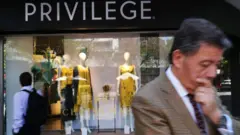 Homem engravatado passa em frente a vitrine de loja com nome 'Privilege'