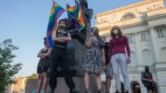 демонстранты на постаменте памятника с радужным флагом
