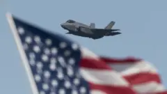 Un avión F35 volando con una bandera de Estados Unidos a la vista