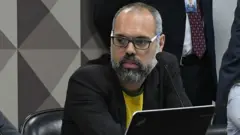 Allan dos Santos sentado diante de microfone na mesa, em sala do Senado