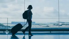 Mulher de perfil arrasta mala em aeroporto, com mochila nas costas