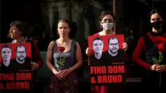 Quatro mulheres, algumas de máscara, aparecem em pé segurando rosas e cartazes com rostos de Bruno e Dom
