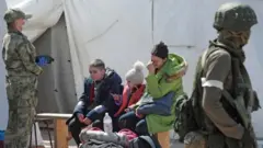 Umugore [urimo kurira] yicaranye n'abana, mu gihe abahungishwa, barimo n'abasivile bavuye mu gace kari hafi y'uruganda rw'ibyuma rwa Azovstal muri Mariupol, bageze mu kigo bagiye kuba bacumbikiwemo by'igihe gito