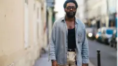 Gali Navangi porte des lunettes de soleil de Port Tanger, une chemise grise vintage, un débardeur noir d'Armani, un pantalon bicolore de Garoze Navangi, une ceinture, le 24 février 2021 à Paris, France.