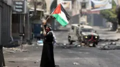 فلسطيني يرفع علم فلسطين أمام عربة عسكرية إسرائيلية أثناء إخراج فلسطينيين من المنطقة