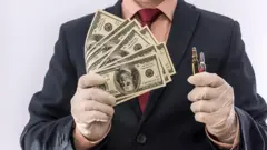 Un hombre con guantes de latex, sosteniendo vacunas en una mano y dinero en la otra