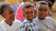 A woman sits with two boys, both of whom wear face paint to mark the occasion.