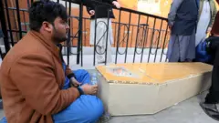 A man sits next to a wooden coffin