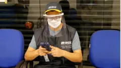 Agente do censo usando boné e colete do IBGE, além de máscara de proteção e face shield contra o coronavírus