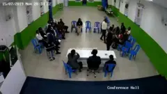 Umugabo (wambaye ishati 'umweru) ava mu kicaro cye agasagarira umukobwa wicaye