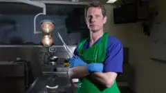 Dr Richard Shepherd dentro de laboratório, com ferramenta em forma de tesoura nas mãos e jaleco