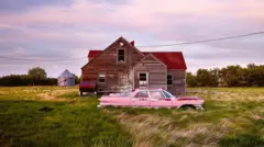 An abandoned pink car in front of a wooden house