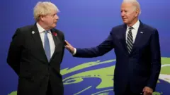 Johnson e Biden em pé, em frente a painel de evento, sorrindo timidamente ao conversarem