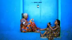 An elderly woman and a girl sit in a blue doorway with a black kitten and a chicken