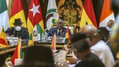 Ecowas and CNSP representatives sit at table surrounded by flags and other emblems of state in Accra, Ghana.