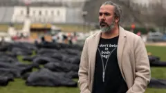 Detrás de Manuel Oliver se ve la instalación de 1.100 bolsas para cadáveres que deletrean las palabras "thoughts and prayers" (pensamientos y oraciones), una frase común de los políticos estadounidenses tras los tiroteos.