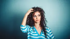 Portrait close-up of a woman wearing a blue and white stripy top, she's looking up, confused, and is standing in front of a blue wall background