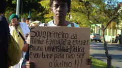 Jovem negro segurando um cartaz que diz 'Sou o primeiro da minha família a entrar em uma universidade pública e vou lutar para não ser o último'