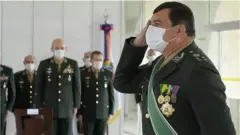 Fotografia colorida mostra em primeiro plano homem branco de meia idade com máscara, cabelo preto curto e uniforme militar batendo continência; ao fundo há outros homens brancos de uniforme militar