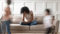 Vultos de duas crianças correndo em volta de sofá, em que mulher está sentada, com cabeça abaixada e apoiada na mão - indicando cansaço