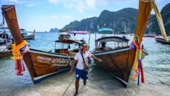 Man in front of long tail boats moored on a beach on Phi Phi Don island in the Andaman Sea.