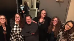 Landon, sua esposa e quatro filhas posam para foto sorrindo, em ambiente domiciliar