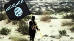 slamic State/Iraq/Syria: A masked Islamic State soldier poses holding the ISIL banner somewhere in the deserts of Iraq or Syria. ISIL publicity image, 2015. (Photo by: Pictures from History/Universal Images Group via Getty Images)