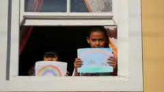 Two children stand at the window holding their drawing of what they miss most - their illustrations show rainbows and ice-cream vans.