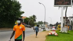 Un homme portant un masque et des gants comme mesure préventive contre le coronavirus COVID-19 passe devant un panneau lumineux portant des messages dans le cadre d'une campagne de sensibilisation au coronavirus COVID-19 dans un quartier résidentiel d'Abidjan le 24 mars 2020.