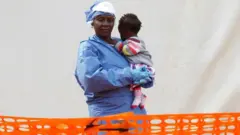 Umukozi w'ubuzima ahagatiye umwana bari ahantu ho kuvurira abarwayi ba Ebola i Butembo muri DR Congo
