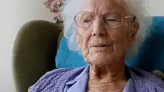 ماي ويليس امرأة مسنة جالسة على كرسي أخضر اللون وعليه غطاء أزرق وهي ترتدي لباس بنفسجي اللون