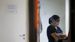 Enfermeira com braços cruzados e máscara olha para baixo dentro de sala de hospital