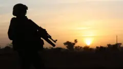 Un soldat monte la garde au coucher du soleil alors que le président français et le président nigérien Mahamadou Issoufou (invisible) participent à une cérémonie militaire au Quartier des Martyrs le 22 décembre 2019, à Niamey, pour rendre hommage aux 71 soldats nigériens massacrés lors d'une attaque le 10 décembre au camp militaire d'Inates, dans la région de Tillaberi, à l'ouest du pays.