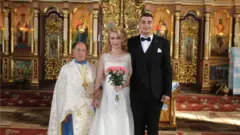 Sladjan and Anne in their wedding day pose with a priest inside a church in Ukraine
