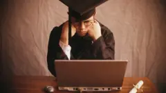 Jovem com beca e chapéu de formatura olha para um notebook com ar frustrado