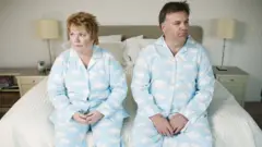 Hombre y mujer vestidos iguales y sentados en el borde de la cama con cara de descontento.