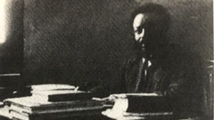 ልሂቅ ገብረክርስቶስ ኮፍ ኢሉ ናብ ካሜራ እናጠመተ፡ ኣብ ጣውላኡ ገለ መጻሕፍቲ ይርአ። እቲ ስእሊ ናይ ቀደም ጻዕዳን ጸሊምን እዩ።