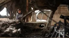 طفل يقف بجانب سيارة محترقة.
