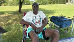 Homem com camiseta branca, shorts azuis e óculos escuros, sentado em uma cadeira de camping sobre a grama