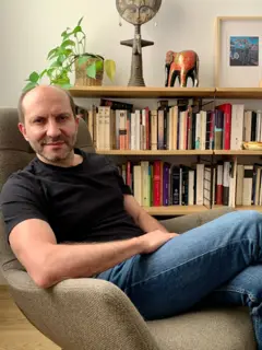 Carlos Granés, está sentado en una silla con jeans y camiseta negra. Detrás tiene una biblioteca con libros.