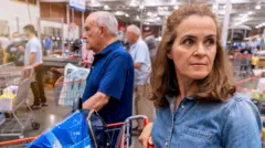 Imagem de uma mulher com cabelos na altura dos ombros, usando uma camisa jeans, parada em frente a um carrinho de compras, com vários consumidores com carrinhos ao fundo