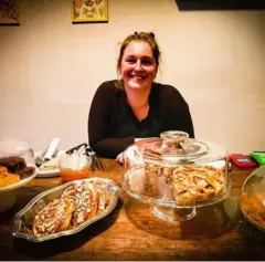 A imagem mostra uma mulher sorrindo atrás do balcão, cercada por doces e tortas expostos em pratos e redomas de vidro. Vestindo roupas escuras e com os cabelos presos, ela apoia os braços sobre a bancada de madeira.