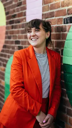 Amanda Mustard sorridente, apoiada em um muro